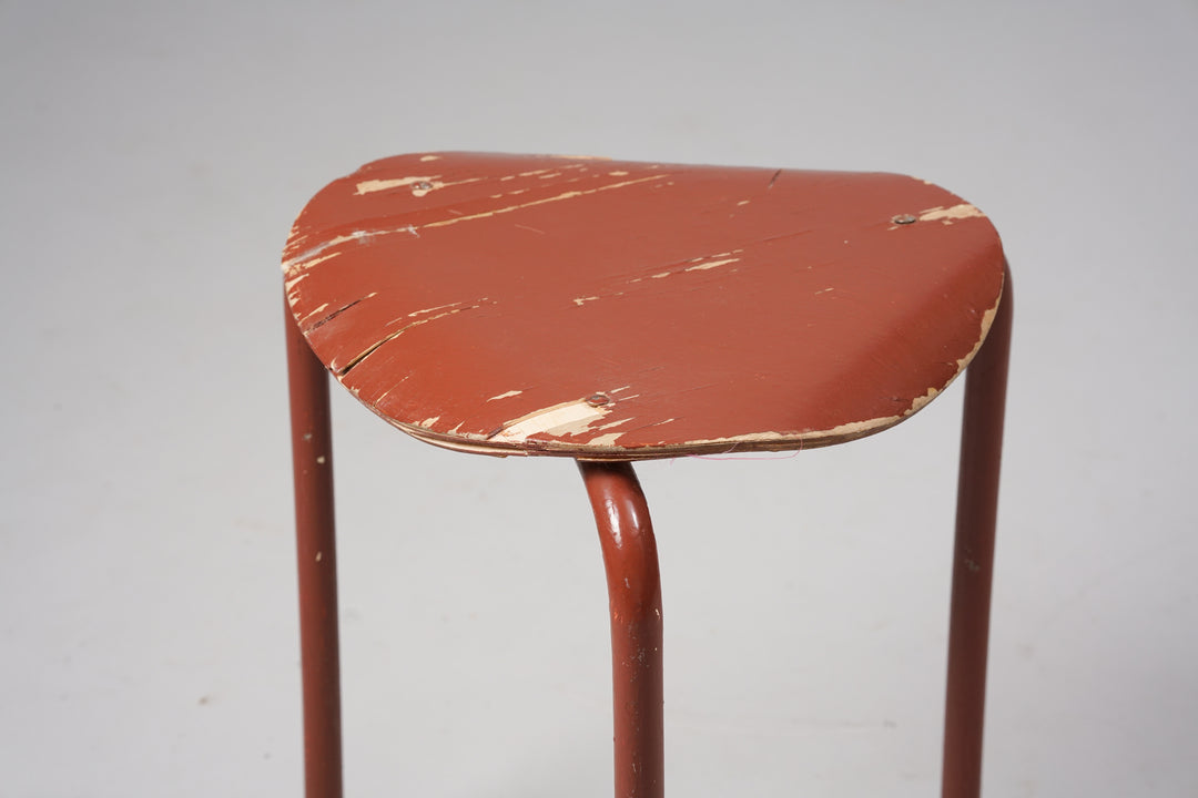 Three legged stool with the edges of the seat bending downwards. The seat is made of plywood and the base of metal. Color of the stool is a brownish red.