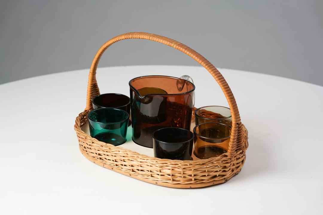 A set that includes six glass cups,a glass ewer and waved basket. The cups are in following colors: Turquoise, black, green, purplish red, yellow and brownish. The ewer is dark orange.