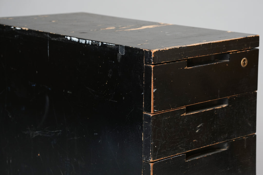 Black wooden filing cabinet with five drawers. The cabinet has a lock on the first drawer and four wheels. Worn from use.