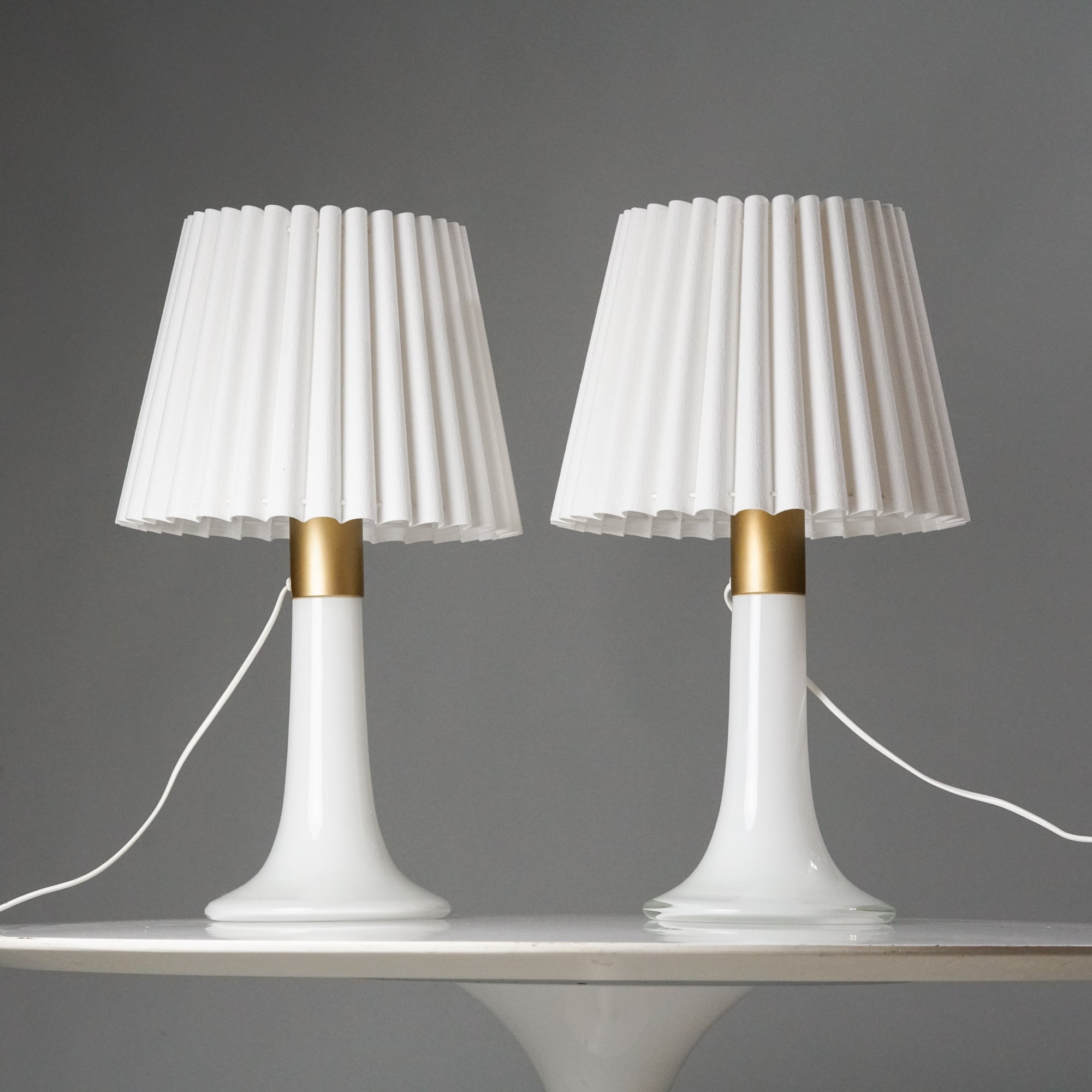 Kaksi samanlaista lamppua, joissa valkoiset laskostetut varjostimet ja valkoisesta lasista tehdyt jalat. Lamppujen jaloissa on maalattu koristelu kullan värillä.