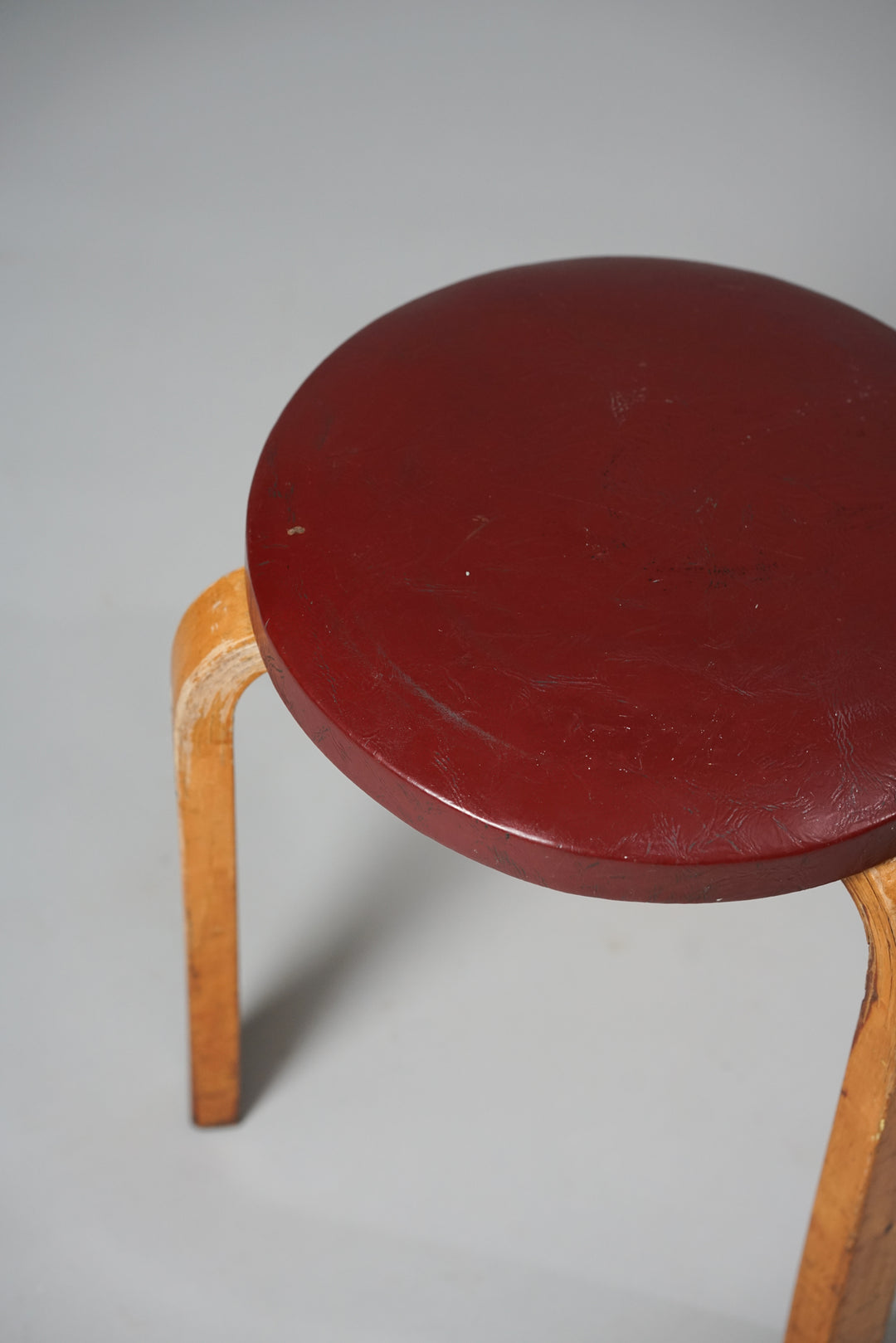 Stool with three wooden legs and dark red round seat. Shows normal wear from use.