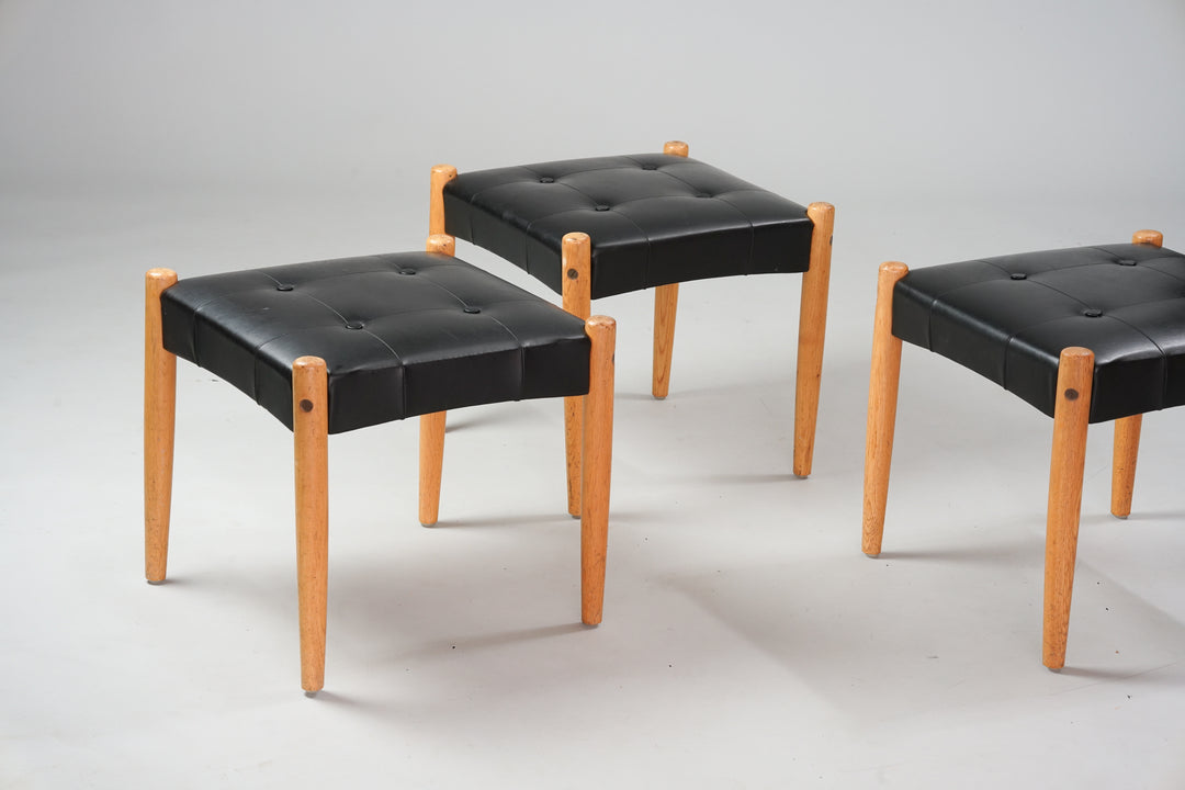 A square footstool with black faux-leather seat and oak legs.