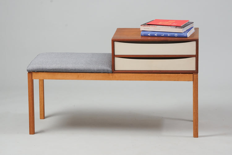 A telephone table with a seat that is upholstered in grey fabric. The frame of the table is birch, and it has a small cabinet part made of teak. There are two drawers that have been painted white.
