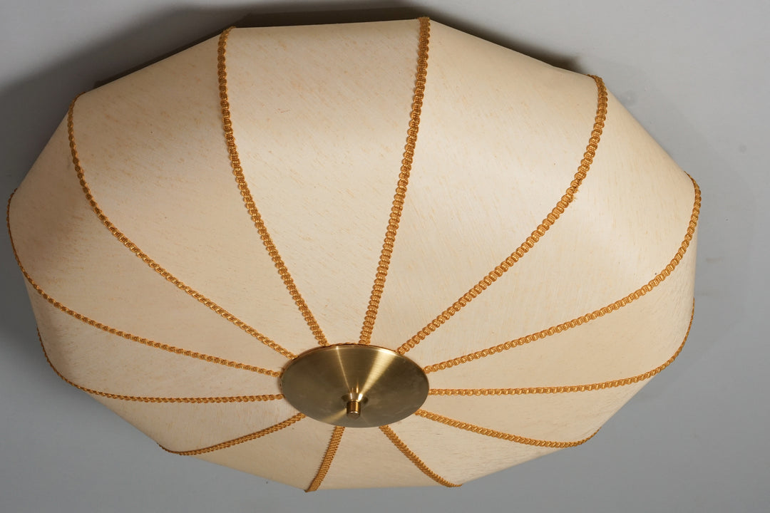 Round flush mount. Beige fabric shade with brown stripes. The stripes come from the sides of the lamp and join in the middle. In the centre is a brass circular plate decoration. 