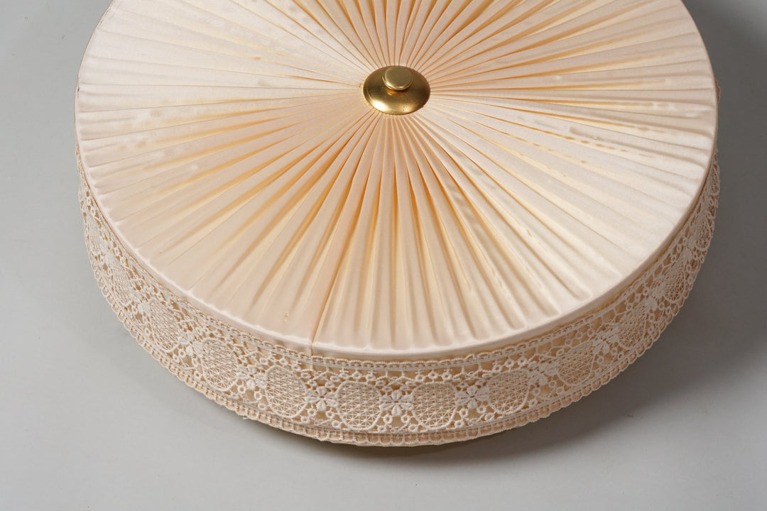 Round flush mount. Pale pink silk fabric, pleated. White lace edging on the side with floral and circular patterns. Round brass detail in the centre.