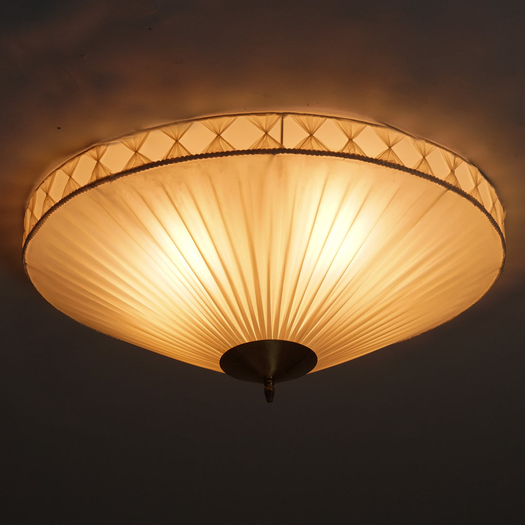 Round flush mount. A natural white plastic shade.The edge of the shade is surrounded by a detail with the same white fabric in vertical bows. In the centre, a circular brass detail. 