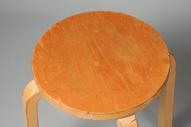 Light brown wooden stool. Four identical legs. Round seat. Seat darker in color than the legs.