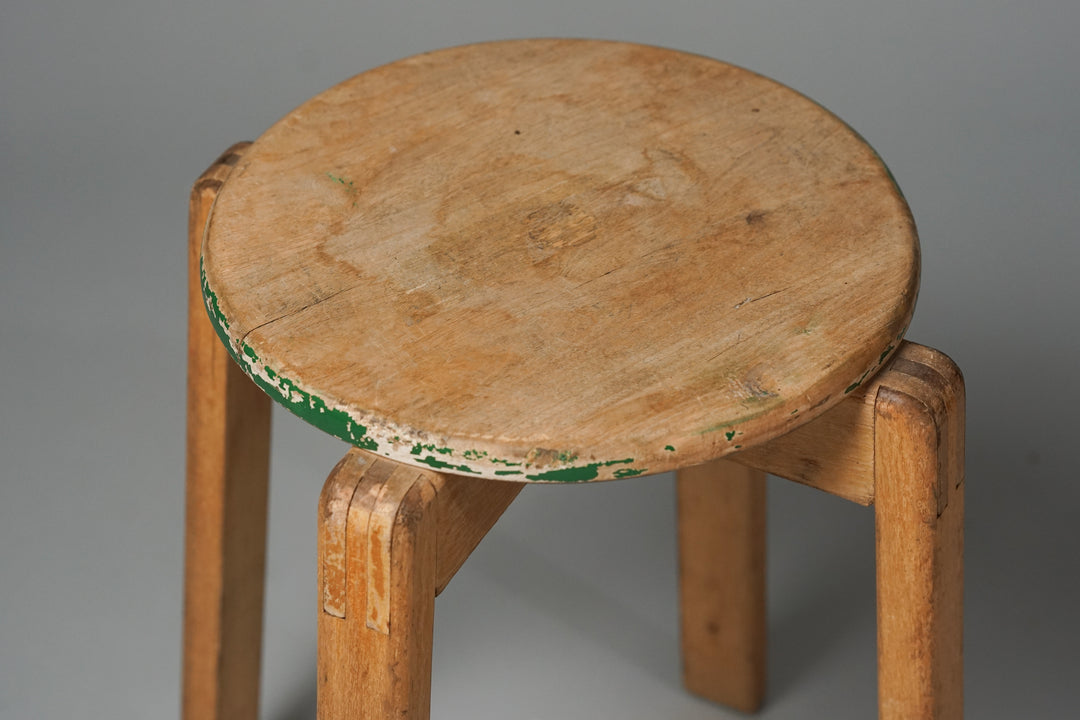 A light brown wooden stool with four identical legs with a wooden joint near the seat. Worn green and white paint on the side of the seat. 