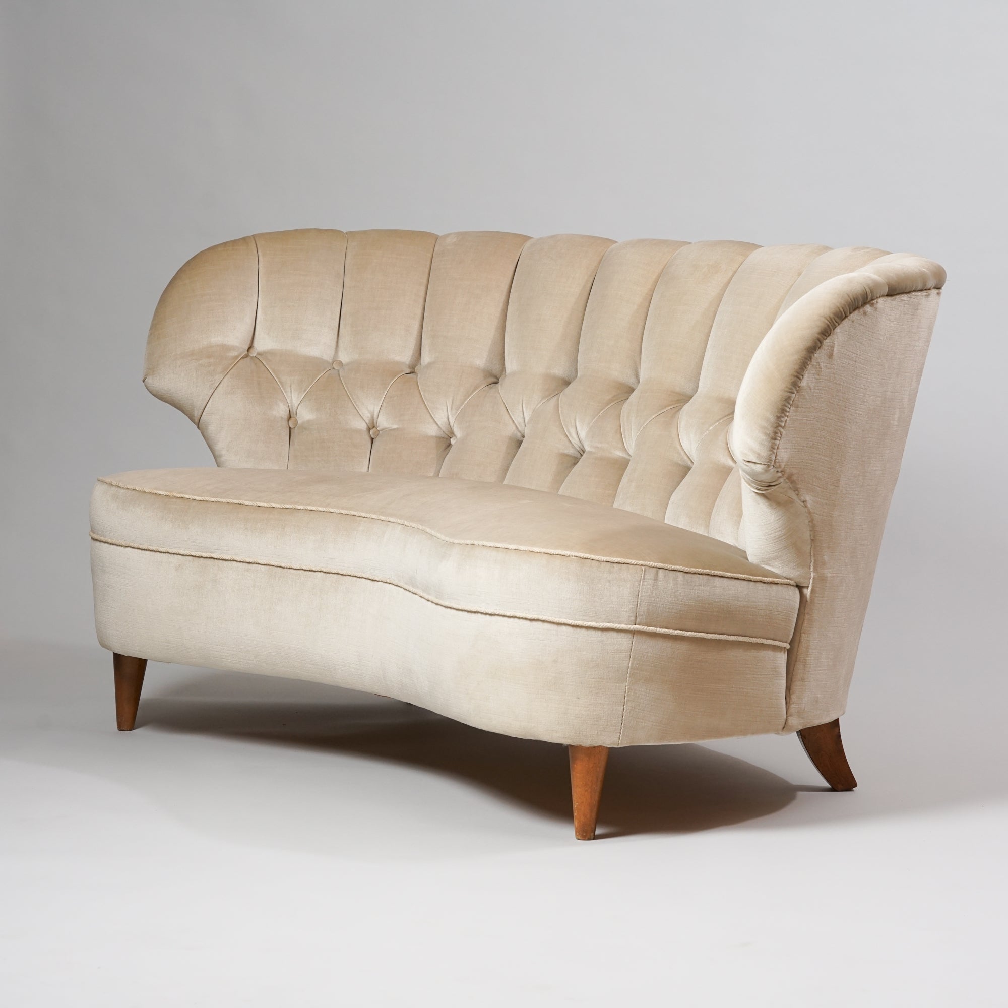 Kaareva sohva, jonka jalat ovat koivua. Sohva on päällystetty kerman väriseen plyysikankaaseen.