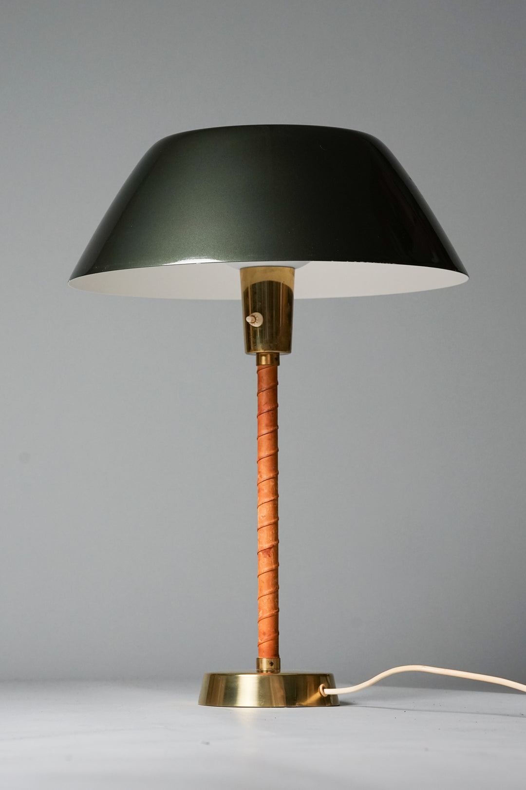 Table lamp with brass base. Red-brown leather trim on the body. Brass detail on the light switch before the shade. Shade in dark green painted metal. 