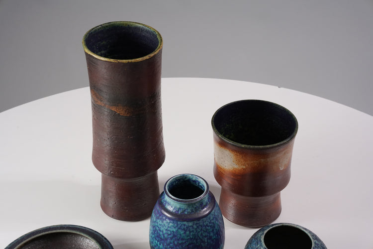 A set of ceramics, with four vases, two bowls and two candlesticks, each holding one candle. Two of the vases are blue, the other two are varying colors of brown. The bowls are brown, while the candlesticks have lighter and darker brown spots.