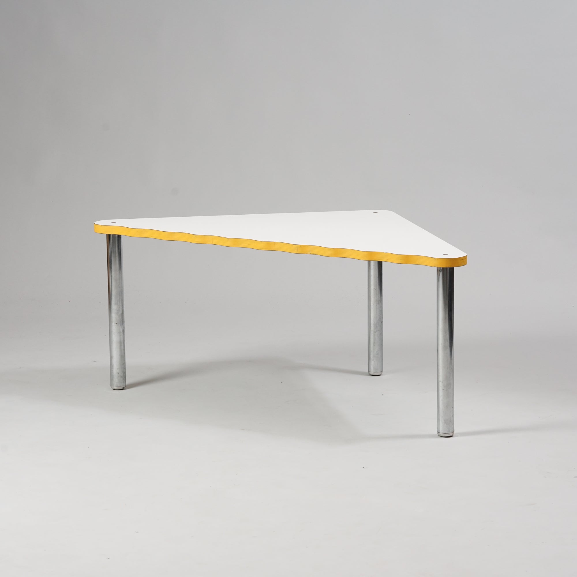 Kolmion muotoinen pöytä, jossa kromatut jalat ja valkoinen linoleumi päällyste. Kolmion pisin sivu on aaltoileva. Pöydän reunat ovat keltaiset.