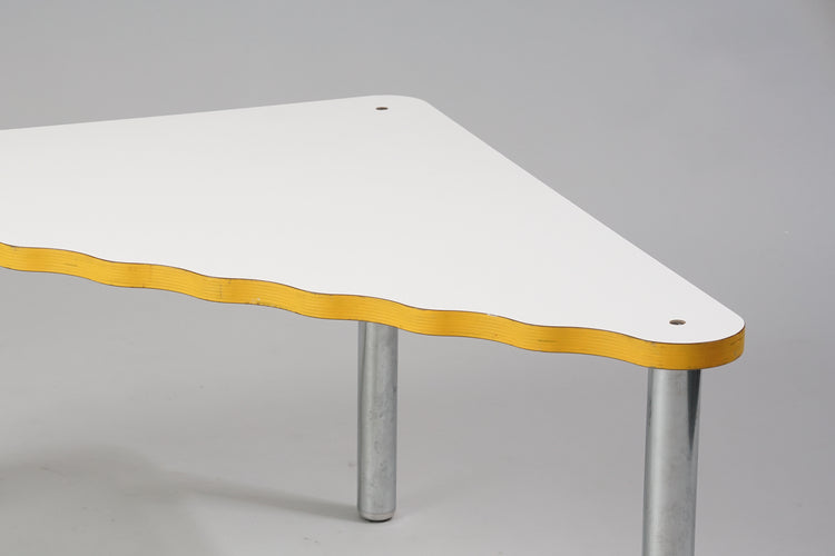Triangle shaped table with chromed legs and white linoleum coating. The longest side of the table has a wavy shape and the edges are yellow.