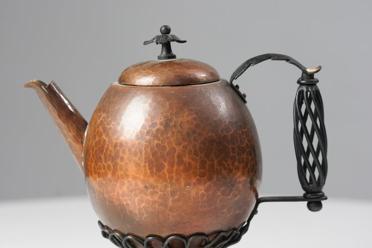 A teapot made of copper with the handle and leg made of decorated wrought iron.