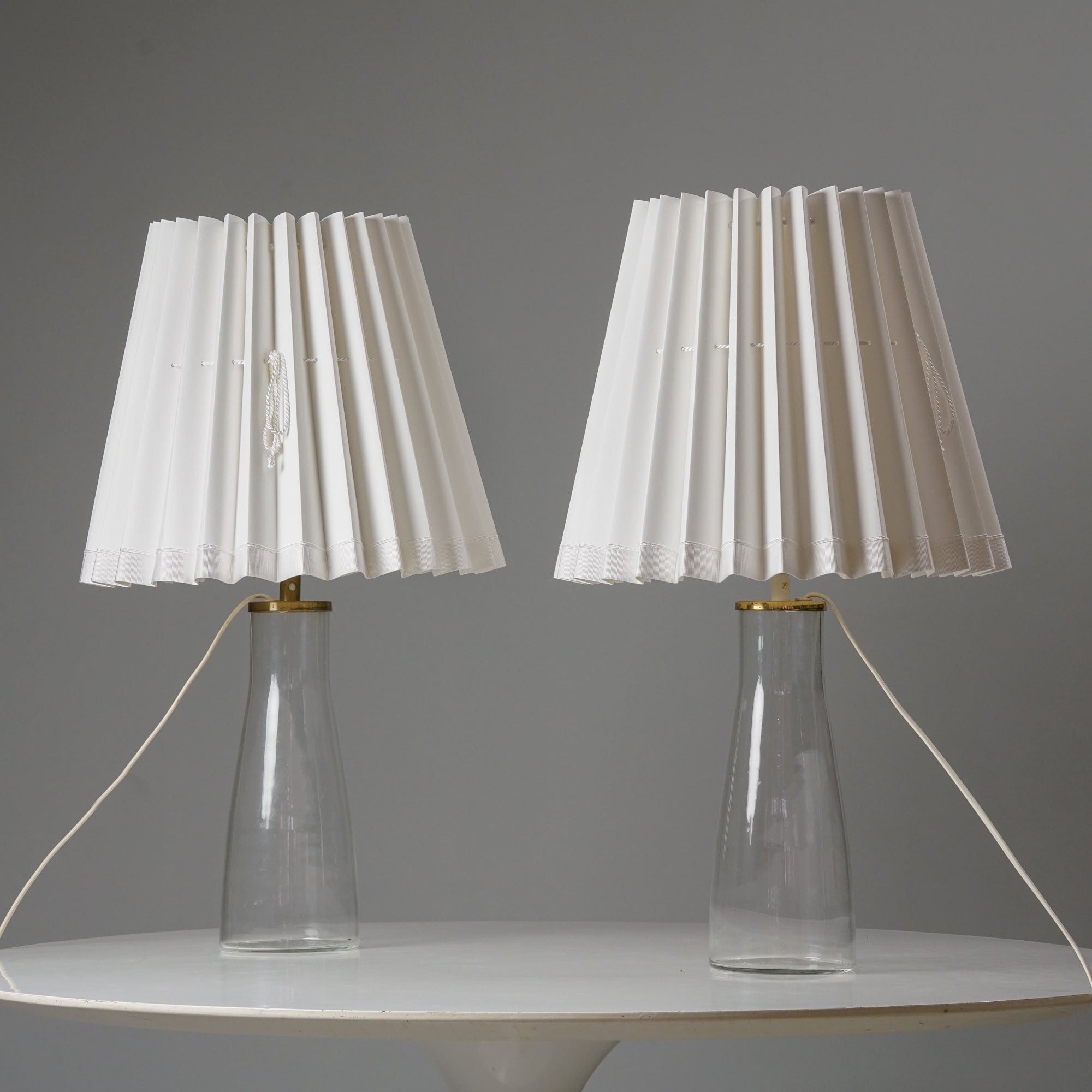 Kaksi samanlaista lamppua. Lampuissa on valkoiset laskostetut varjostimet ja läpinäkyvät lasiset jalat messinkikoristelulla. 