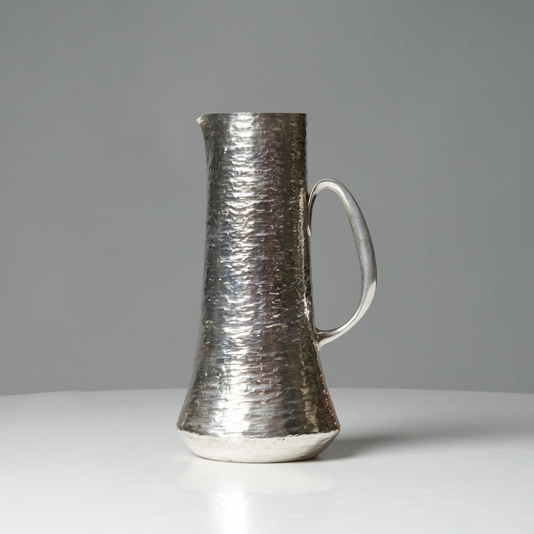 Jug made of silver with a pouring lip and handle. The texture of the jug is uneven.