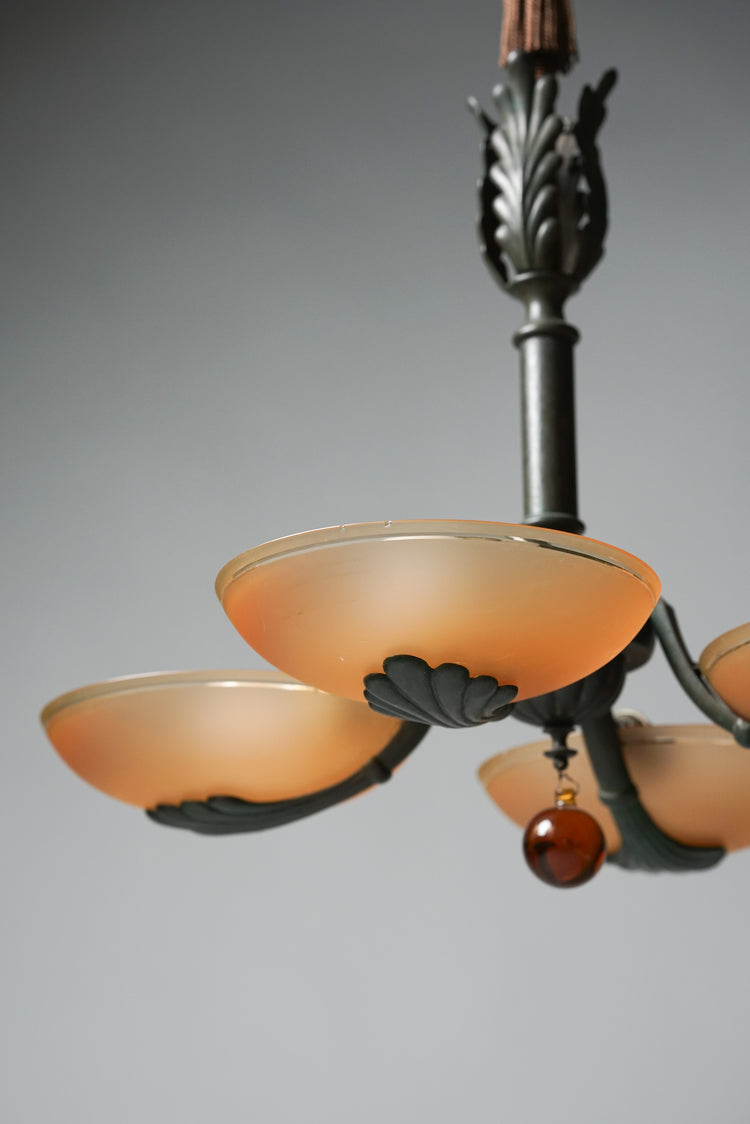 Lamp made of wrought iron with four lamps. The domes of the lamps are orange opal glass. The frame of the lamp is decorated with lacing and has a brown glass ball hanging off it.