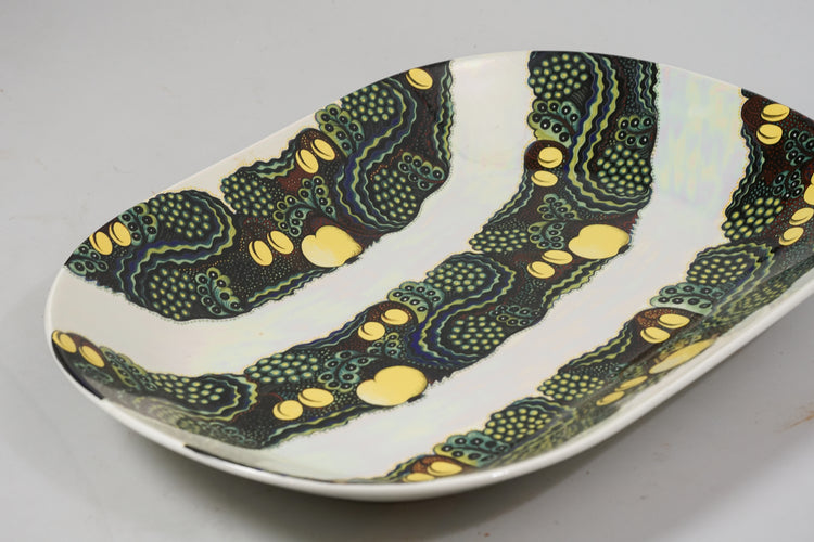 The background of the decorative plate is white with three green parts going through it. The green parts are covered in leaves and have yellow fruits on them.