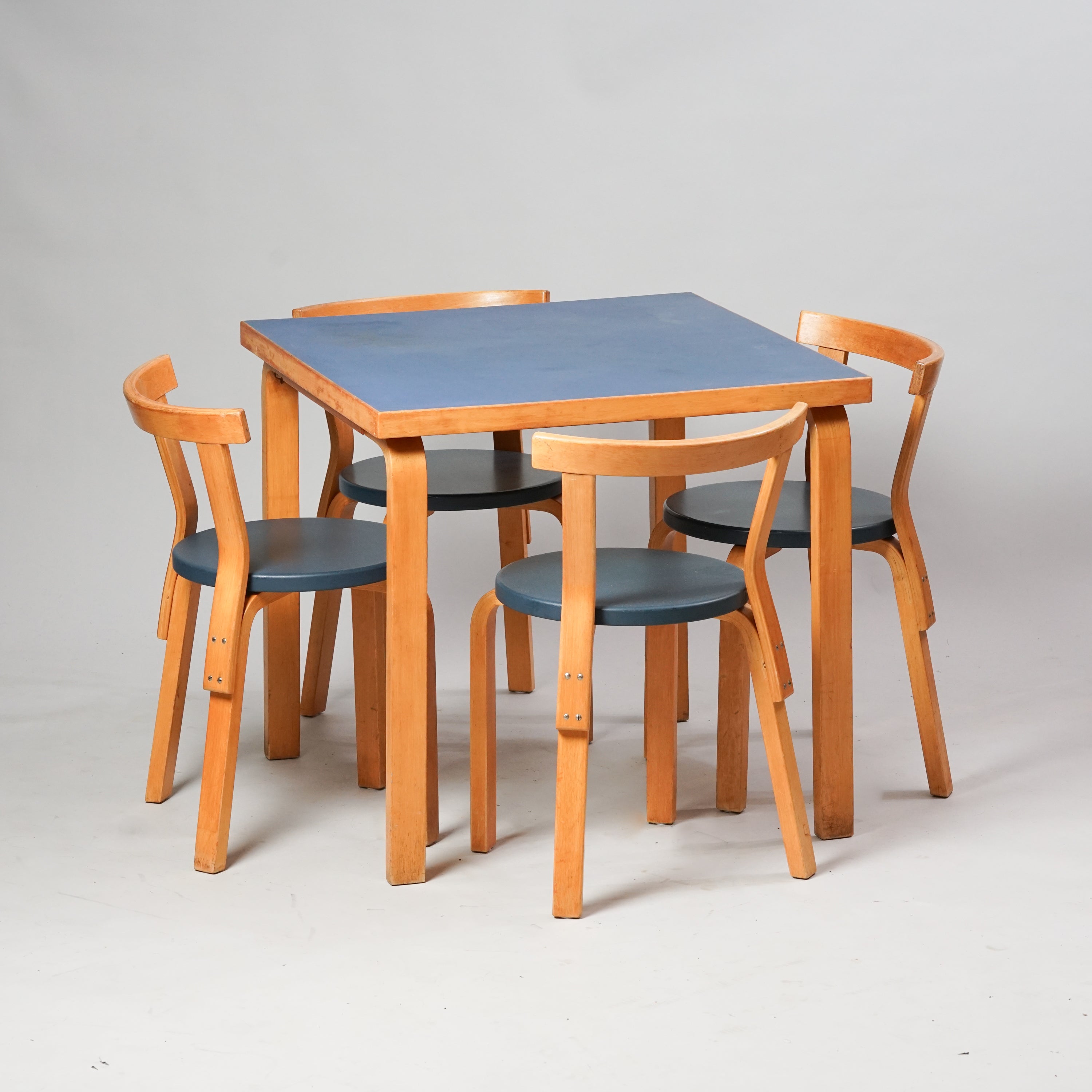 Neliönmuotoinen puupöytä, jonka työtaso on sininen. Neljä tuolia ovat kaikki samanlaisia. Tuolit ovat tehty puusta, ja niiden pyöreät istuimet ovat väriltään harmahtavan siniset.