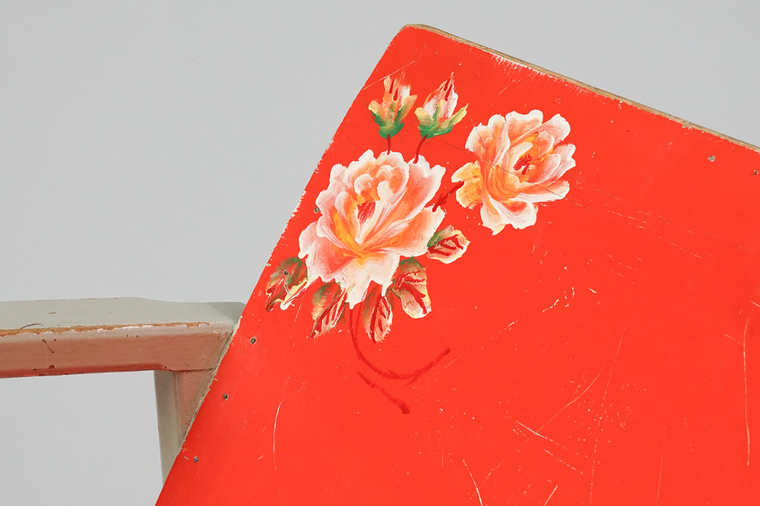 Small rocking chair where the seat and backrest are red. In the top corner of the backrest are painted flowers. Otherwise the rocking chair is a white color.