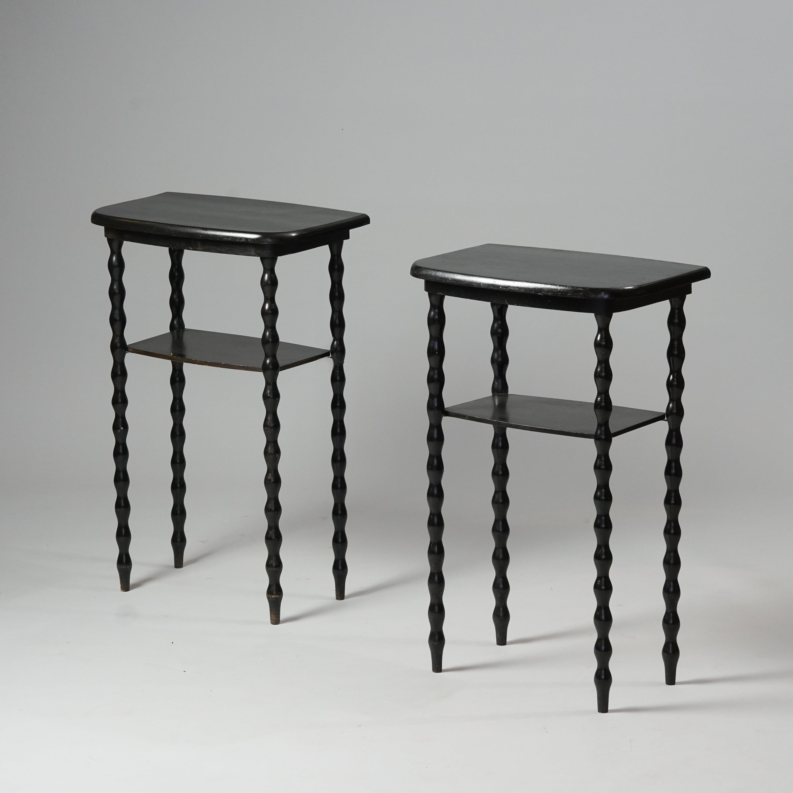 Kaksi samanlaista sivupöytää. Puinen pöytä on maalattu mustaksi ja siinä hylly tason alla. Sivupöydän jalat ovat funky-tyylistä aaltoilevalla muodollaan.