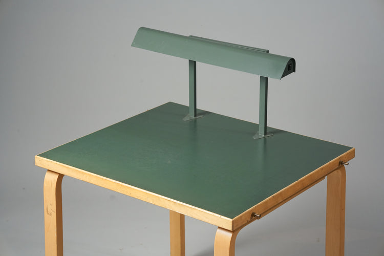 A desk made of birch with the desktop being green in color. The desk has a lamp attached to it that is green as well.