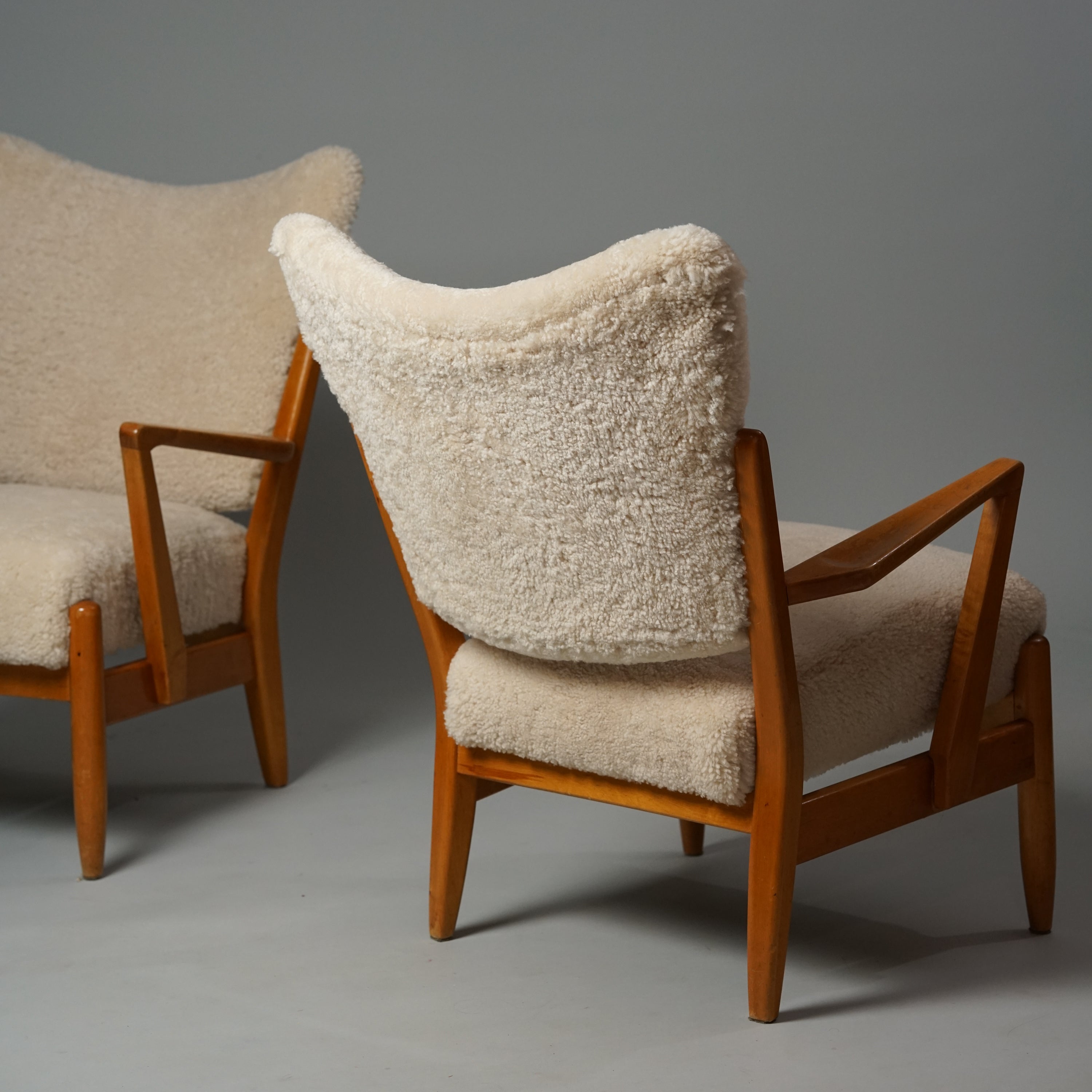 Kaksi samanlaista wingback tuolia, joiden runko on koivua. Istuin ja selkänoja on verhoiltuvaalealla lampaanvillalla.