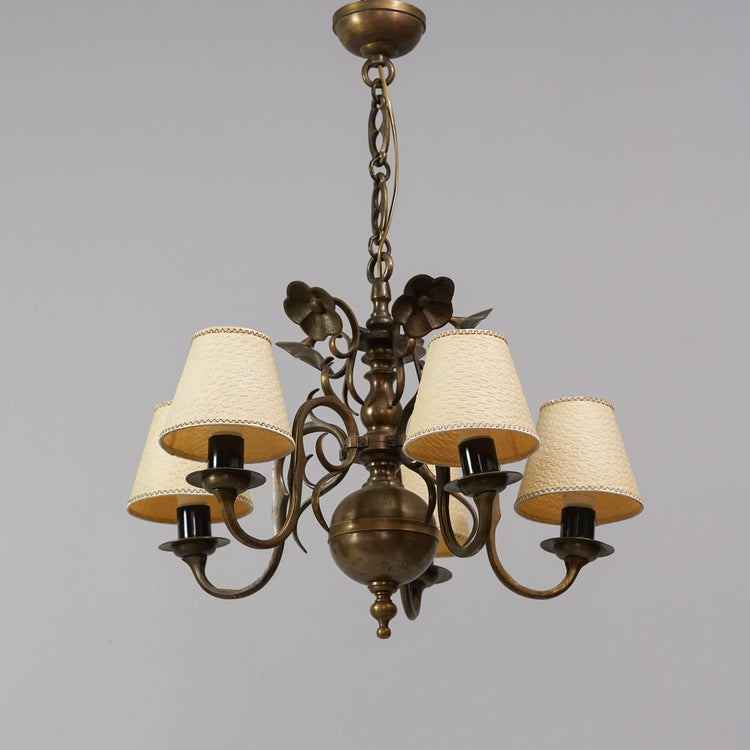 A brass chandelier with flower decorations. The lamp has five lights and they have off-white paper shades. 
