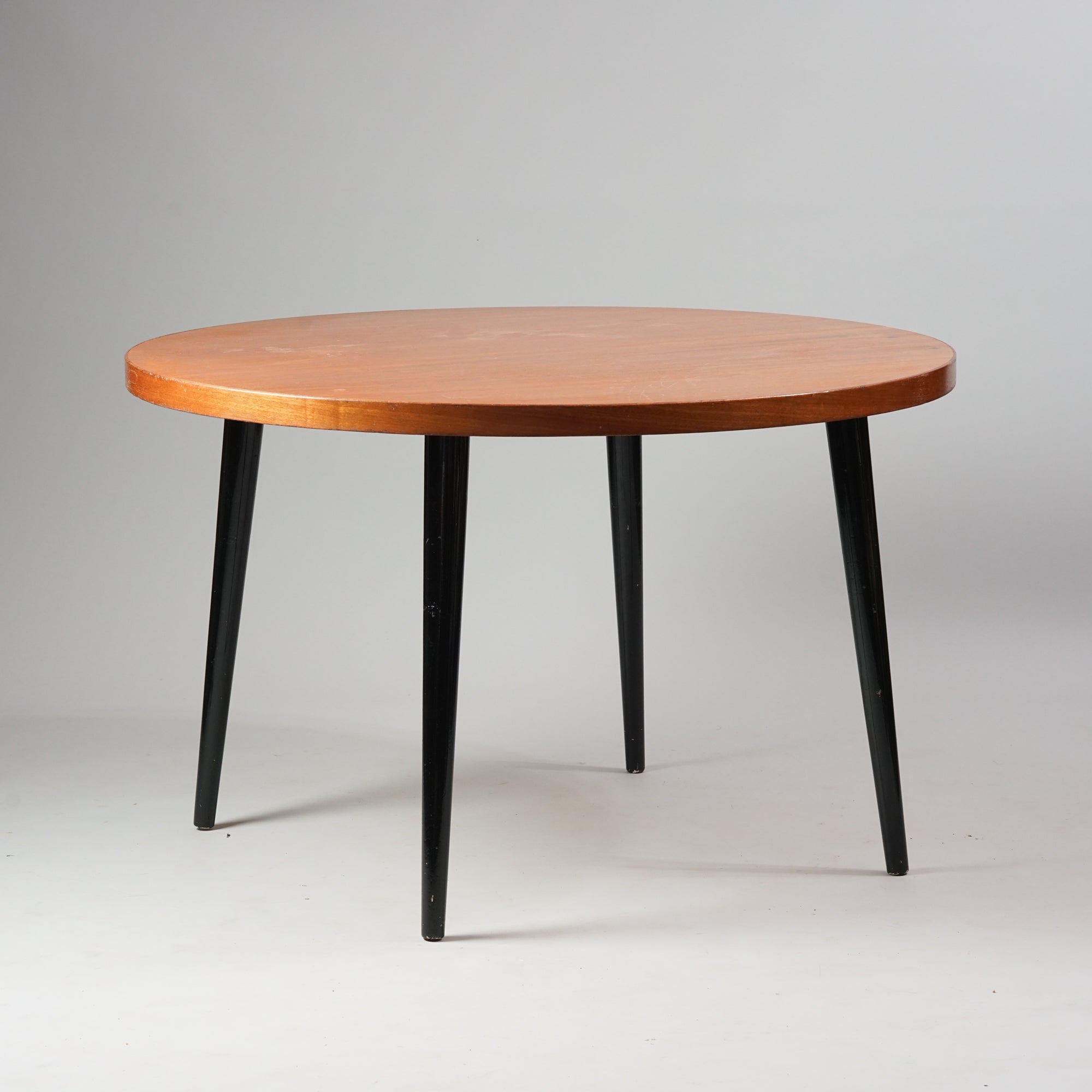 Pyöreä pöytä, jonka taso on tiikkiä ja jalat mustaksi maalattua puuta. 
