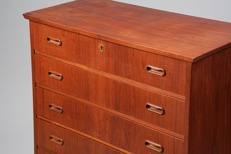 A chest of drawers made of teak. There are five drawers and the top one has a lock.