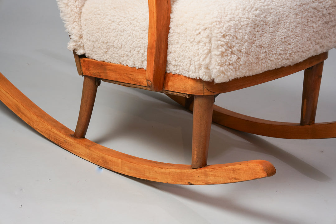 A rocking chair with legs and armrests made of wood. The seat and backrest are upholstered in white sheep's wool.