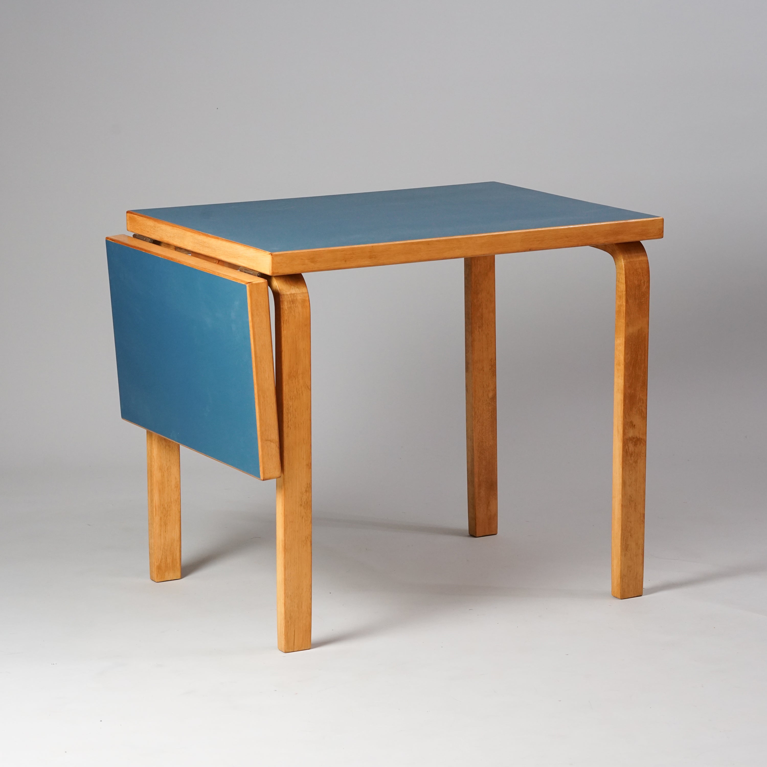 Pöytä, jossa toisessa päässä taitettava osa. Työtaso on väriltään sininen, muuten pöytä on puun värinen.
