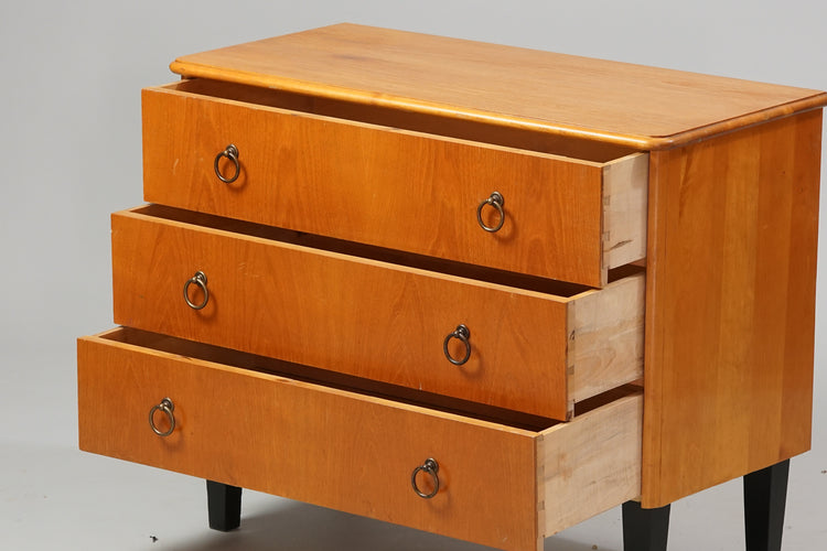 Chest of drawers made of birch and elm, with three drawers. The drawers have brass handles, and the legs are black.