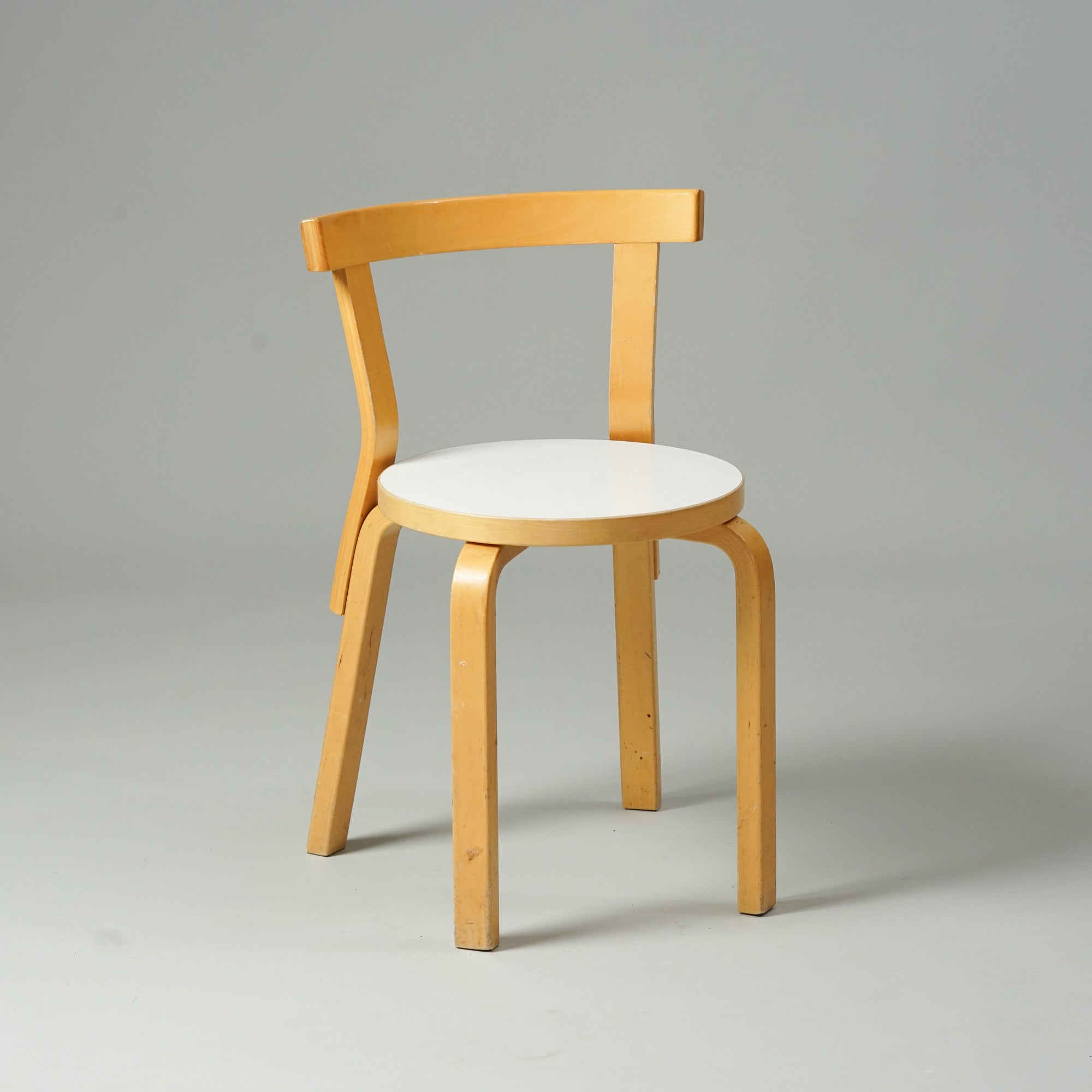 Koivusta tehty tuoli, jossa pyöreä istuin valkoisella linoleum päällisellä. Tuolissa on ohut selkänoja ja neljä jalkaa.