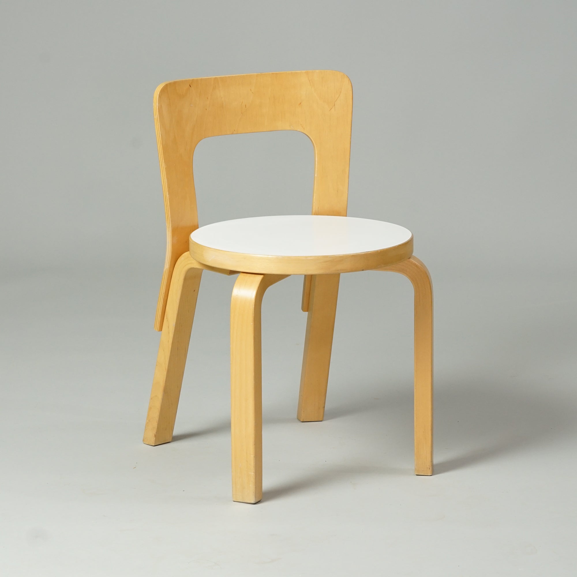 Koivusta tehty tuoli, jossa pyöreä istuin valkoisella linoleum päällisellä. Tuolissa on selkänoja ja neljä jalkaa.