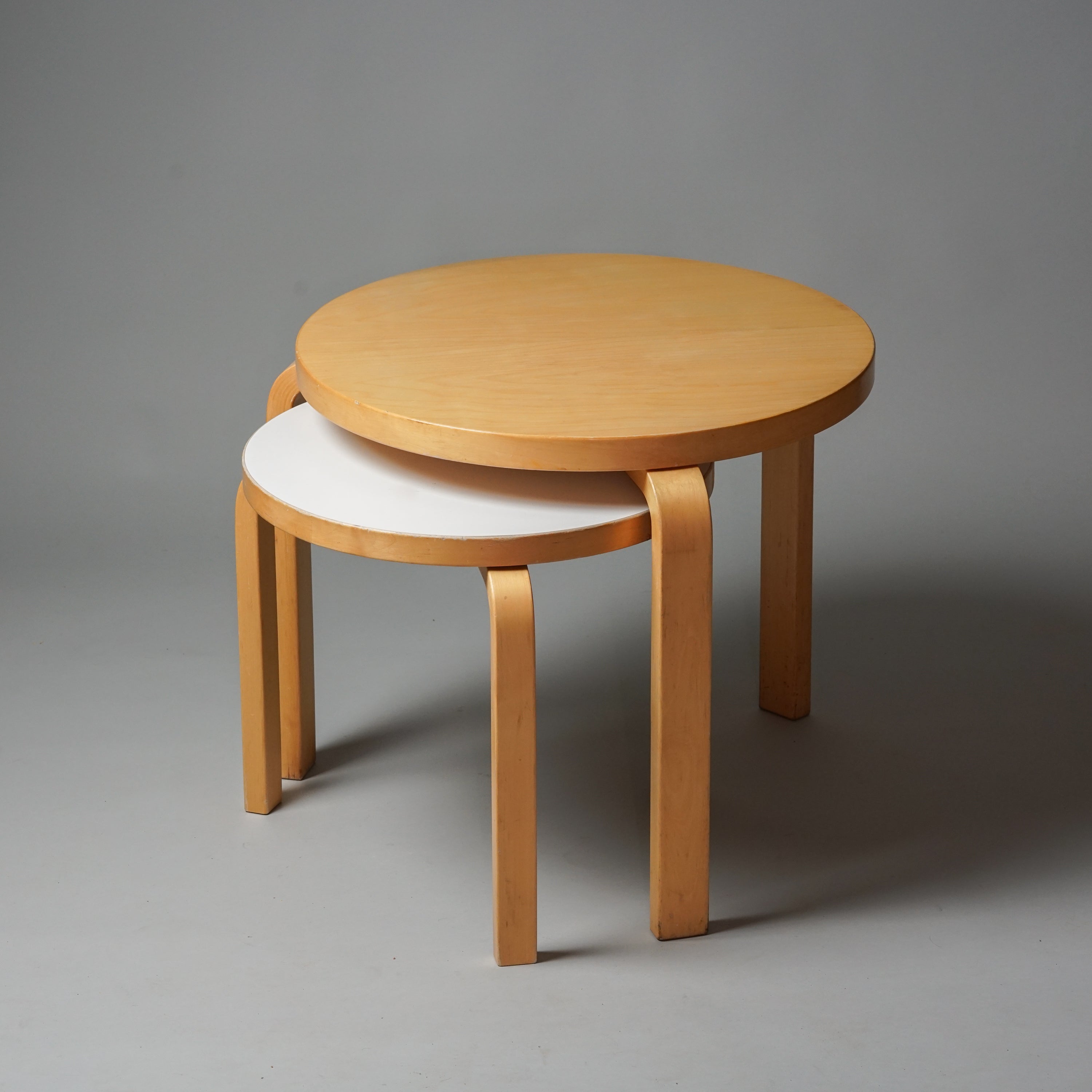 Kaksi pyöreää pöytää, jotka ovat tehty koivusta ja joissa on kolme jalkaa. Pienemmässä pöydässä on valkoinen linoleumi päällinen. 