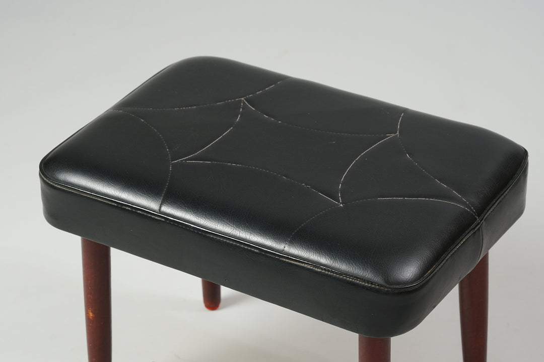 Stool with dark teak legs and black faux leather seat with a surface pattern.