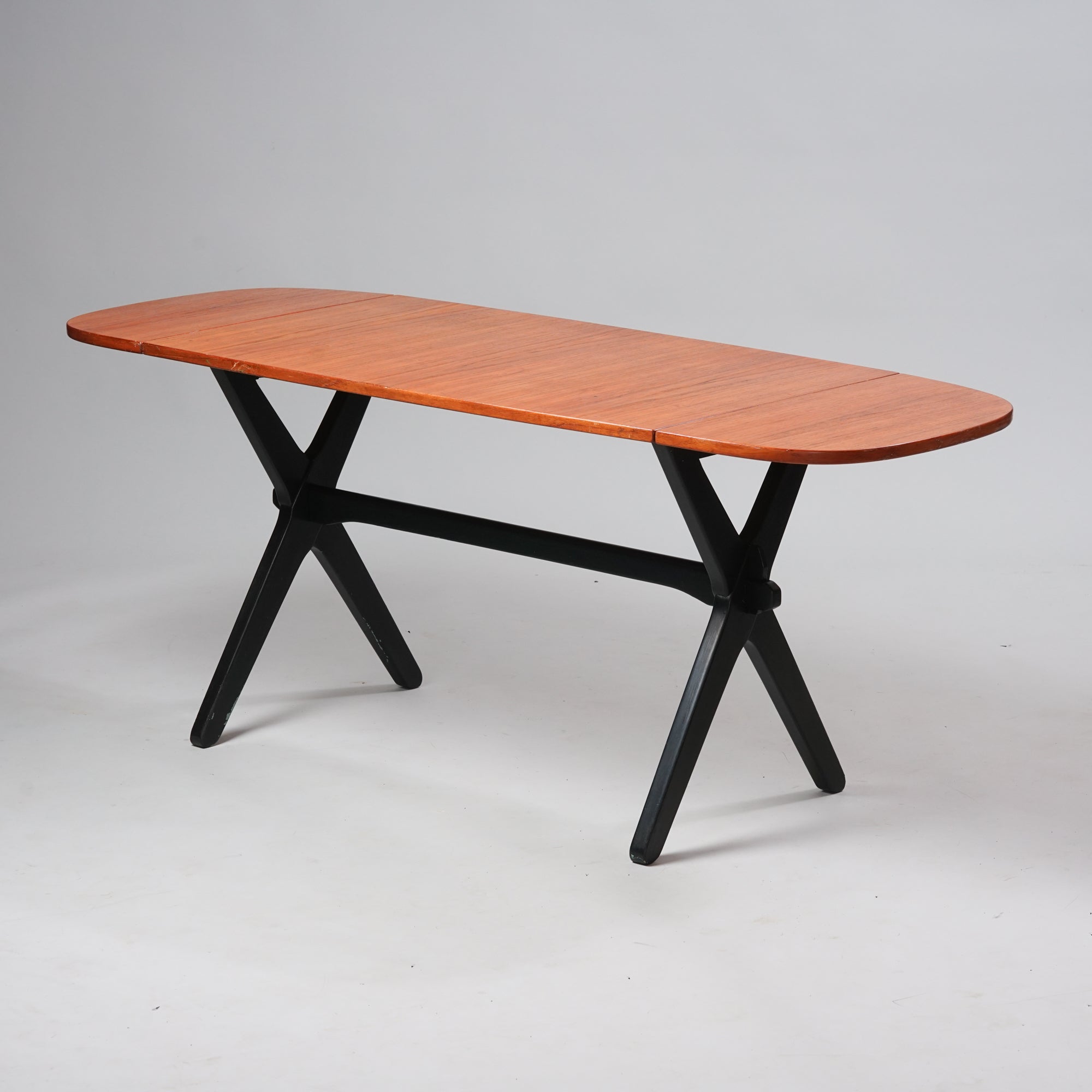 Tiikkitasoinen pöytä, jossa molemmissa päissä on taitettava lisäosa. Pöydän puiset jalat ovat maalattu mustaksi.