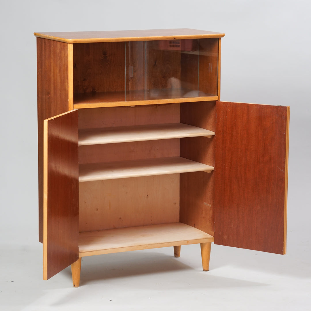 A showcase cabinet made of teak and birch. Has two doors, one has a keyhole and inside are two shelves. The top part of the cabinet has two sliding glass doors. 