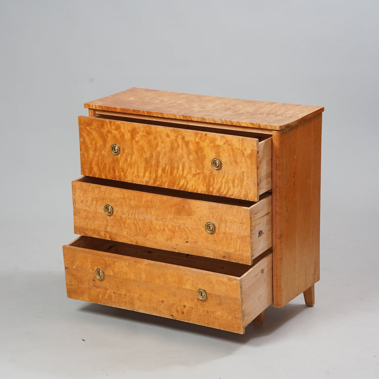 A chest of drawers made of birch and flamed birch. There are three drawers which all have two brass handles.