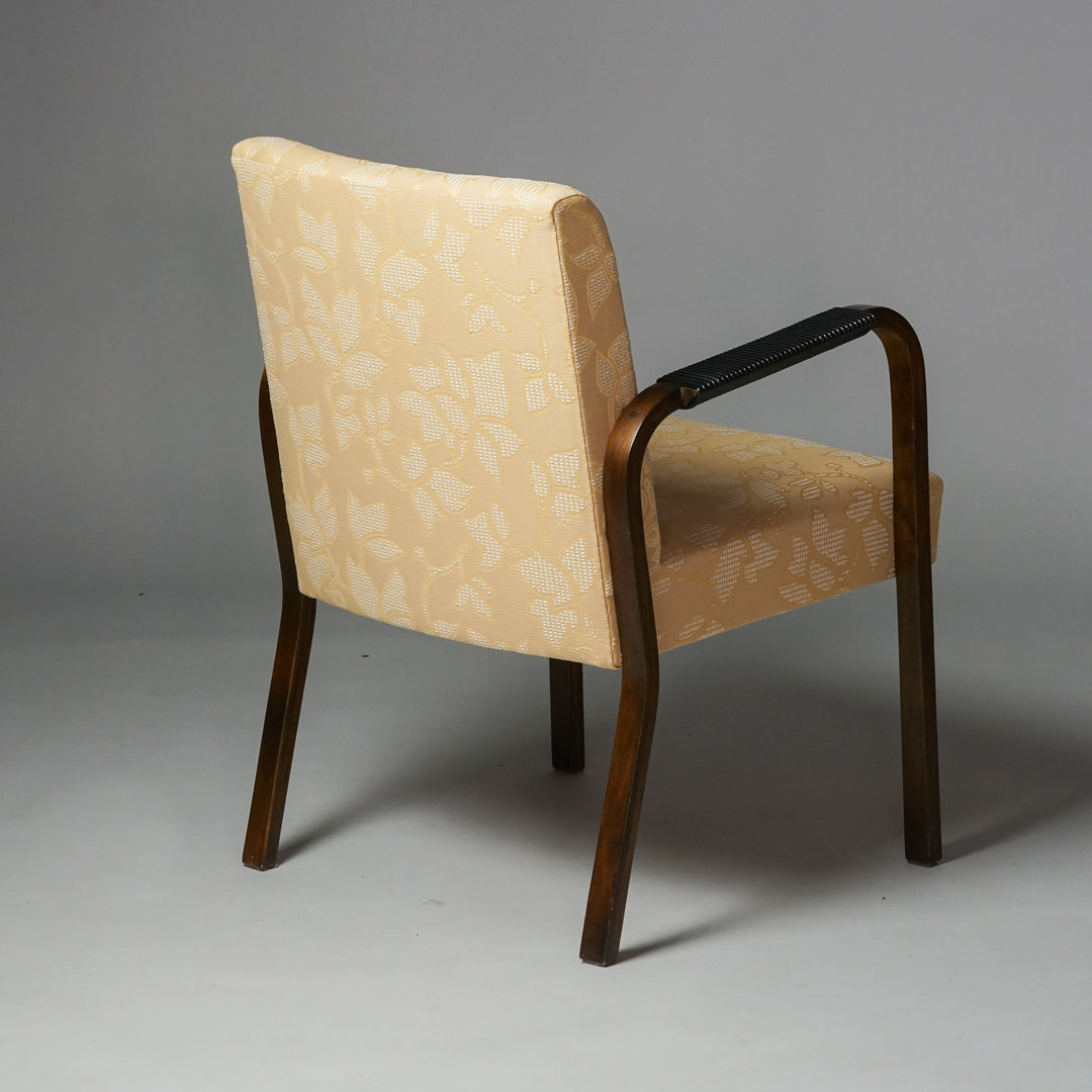 Armchair with deep brown armrests. The armrests have black plastic wrapping details. The armchair is upholstered with yellow fabric that has flower details. 