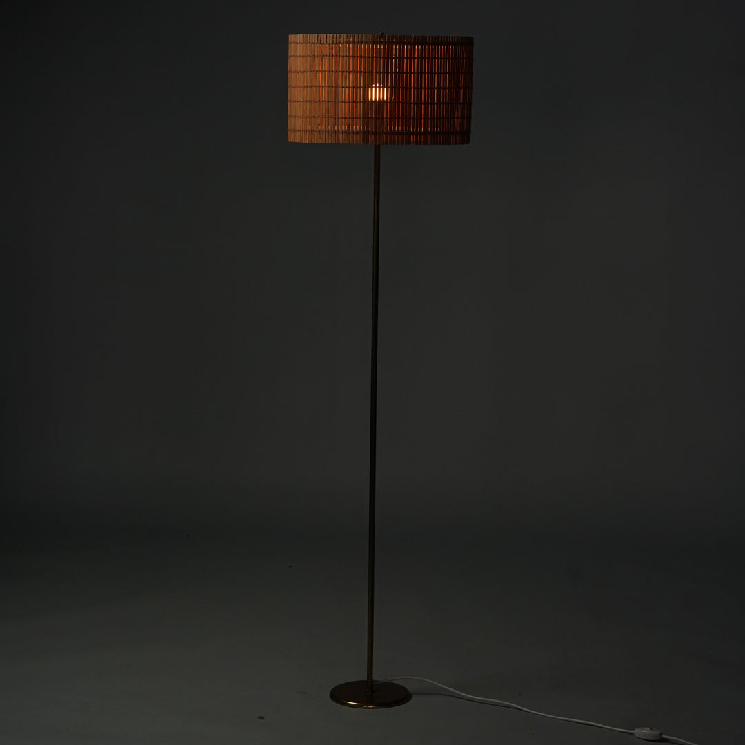 A floor light with brass frame. The lampshade is round-shaped and its made of wood slats.