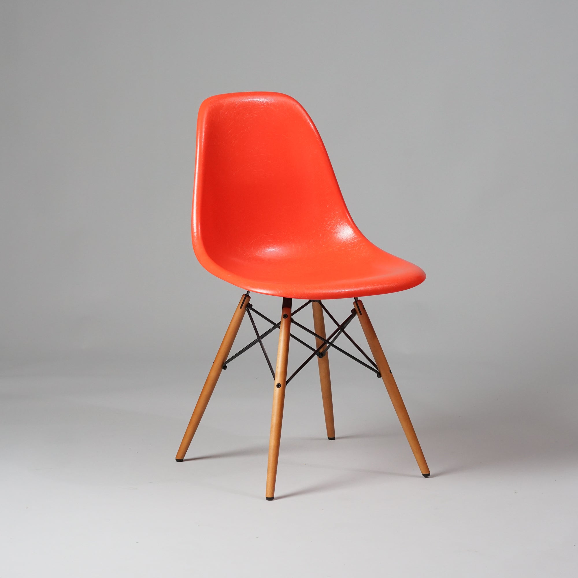 Tuoli, jonka jalat ovat vaahteraa ja jalkojen välissä olevat tangot metallia. Punertava oranssi istuinosa on lasikuitua.