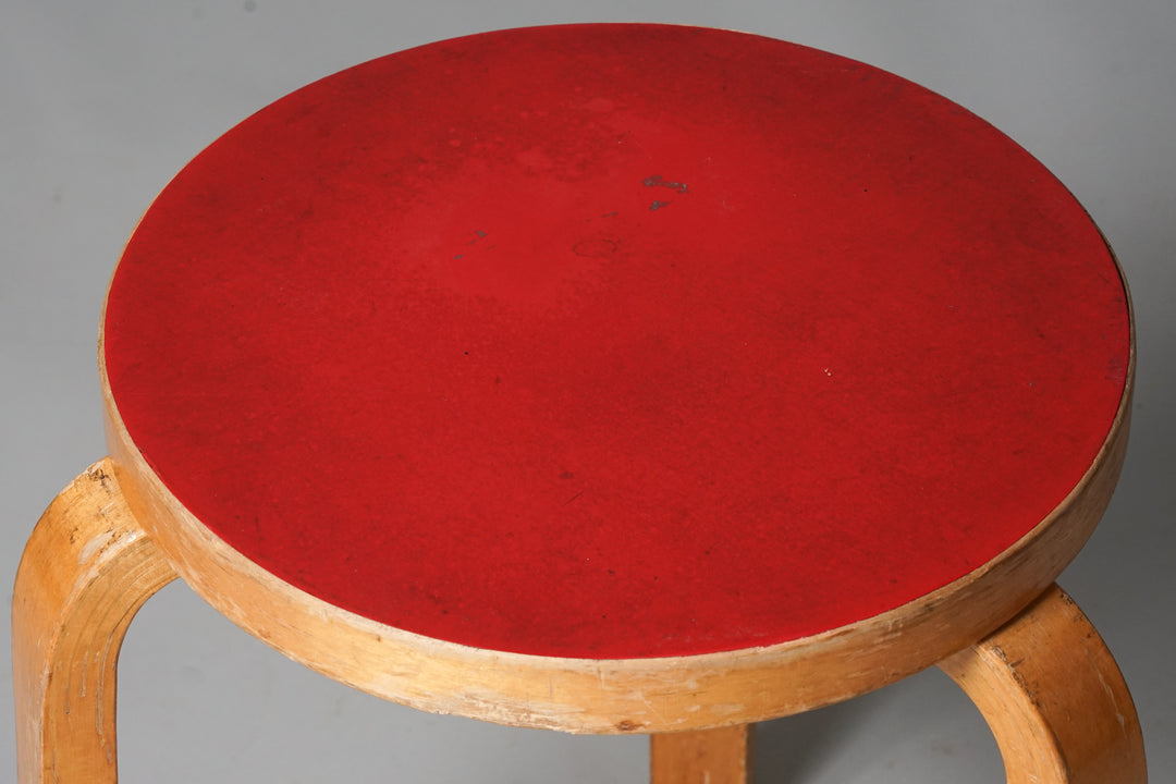 Round stool with red seat and three honey-coloured birch legs.