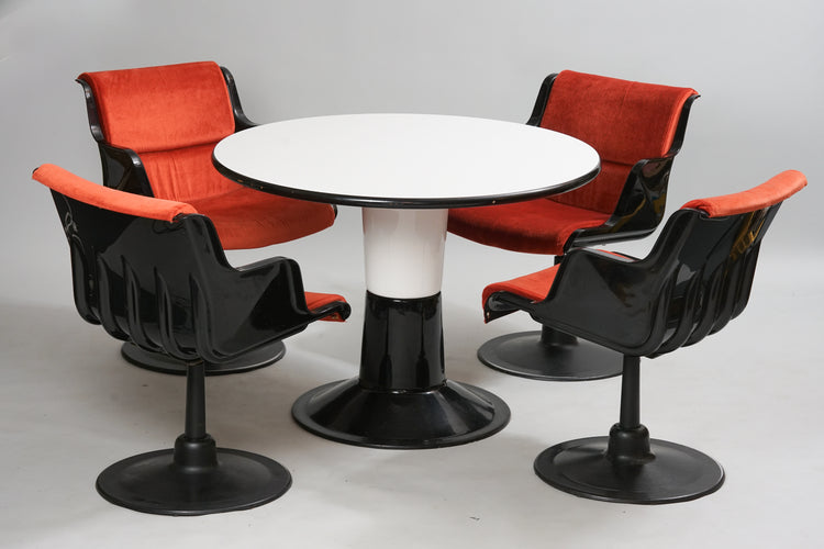 Four identical chairs and a round table. Chairs with plastic seat, metal tube frame with round iron plate at the bottom. The chairs swivel. The chairs are black, the seat orange. The table top is white with a black border. Table leg in metal. Round, half black and half white.