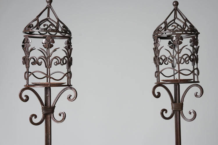 Two identical long candlesticks. The candle is placed in a birdcage-like decorative cage, which top opens. The cage contains floral ornaments. The candle bottom and the details at the bottom of the cage are circular.