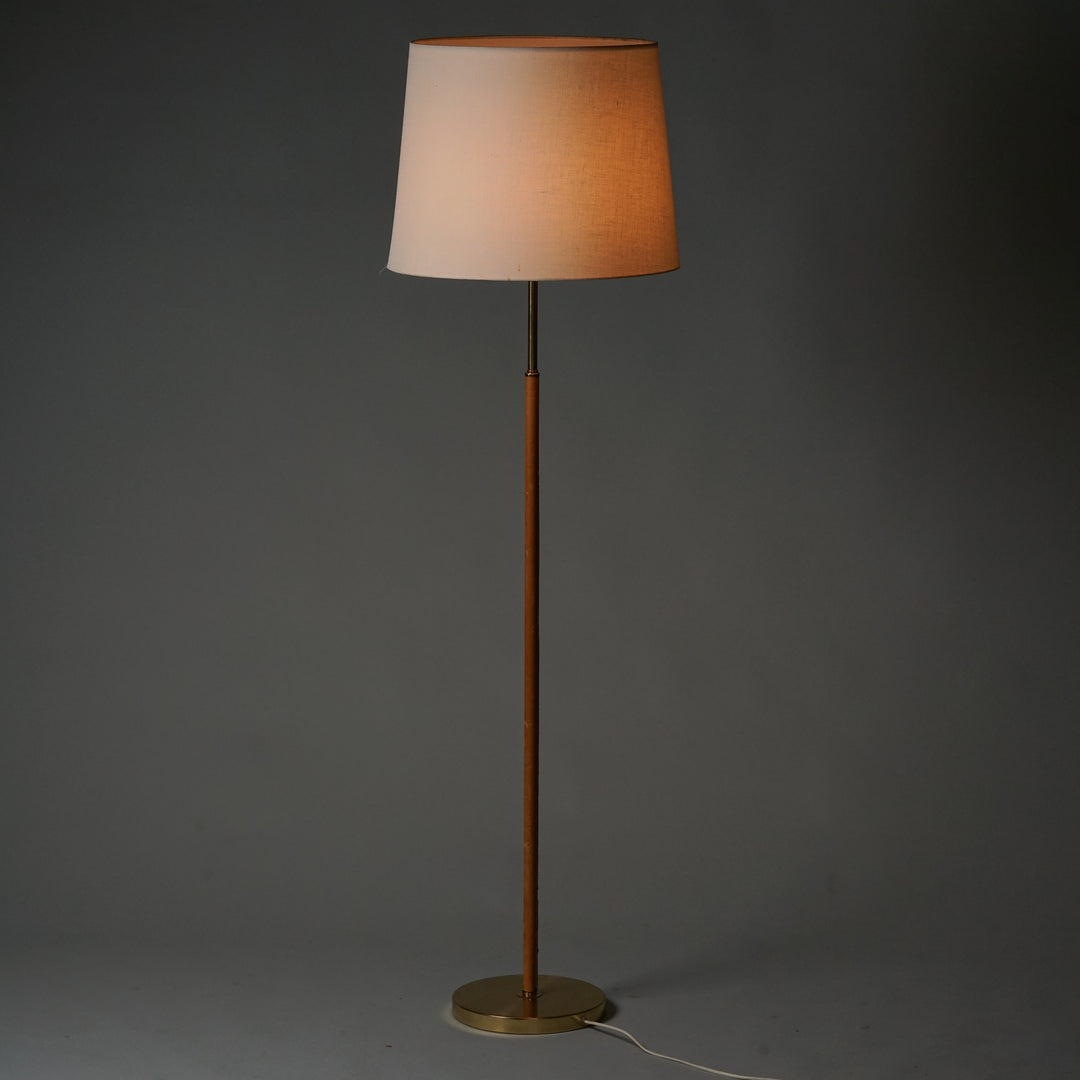 Two identical floor lamps with round brass base. Orange brown leather wrapping and brass on the frame.The lampshade is round and made of white fabric. Inside the shade is a white acrylic frame that holds the shade in place.