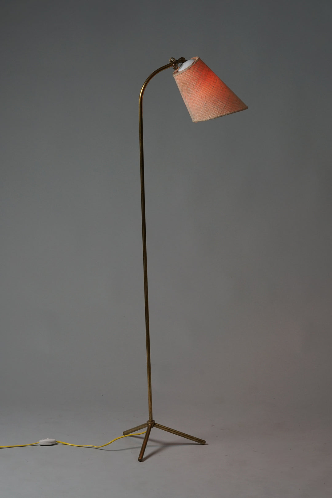 A floor lamp with a slim brass body and three support legs. Lampshade in beige fabric with textured surface. At the end of the shade a small brass ringlet detail. The light switch is attached to the cord on the floor.