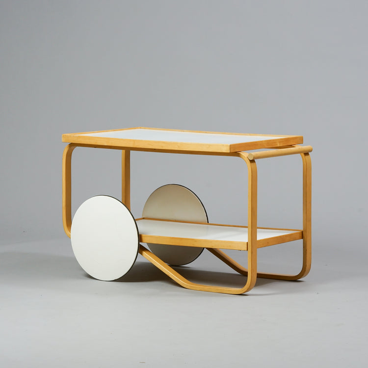 A serving trolley made of birch with two white laminate tops. Has two big white wheels.