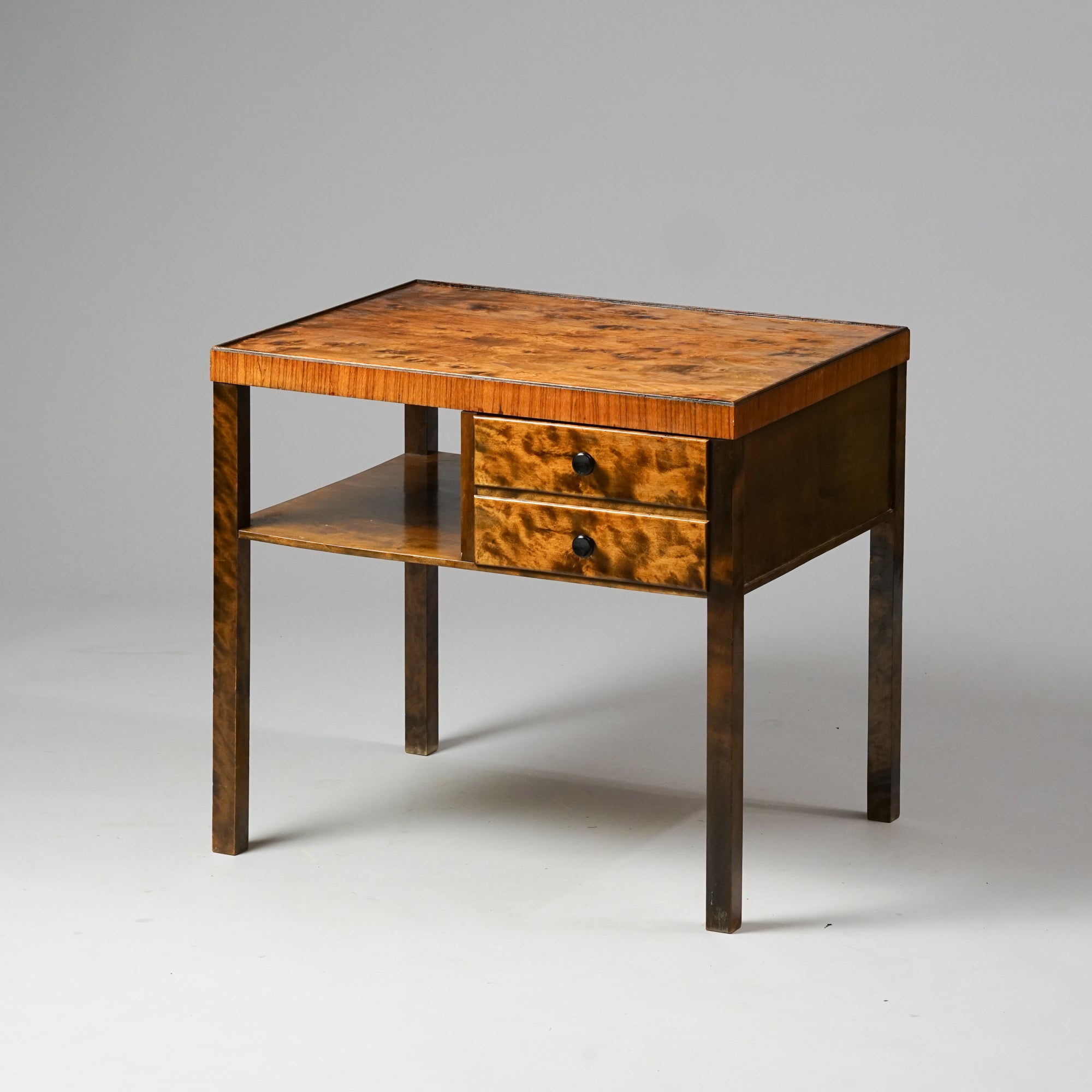 Koivusta ja loimukoivusta tehty pöytä, jonka pinnassa on elävä kuviointi. Pöydässä on kaksi vetolaatikkoa ja yksi hylly. 